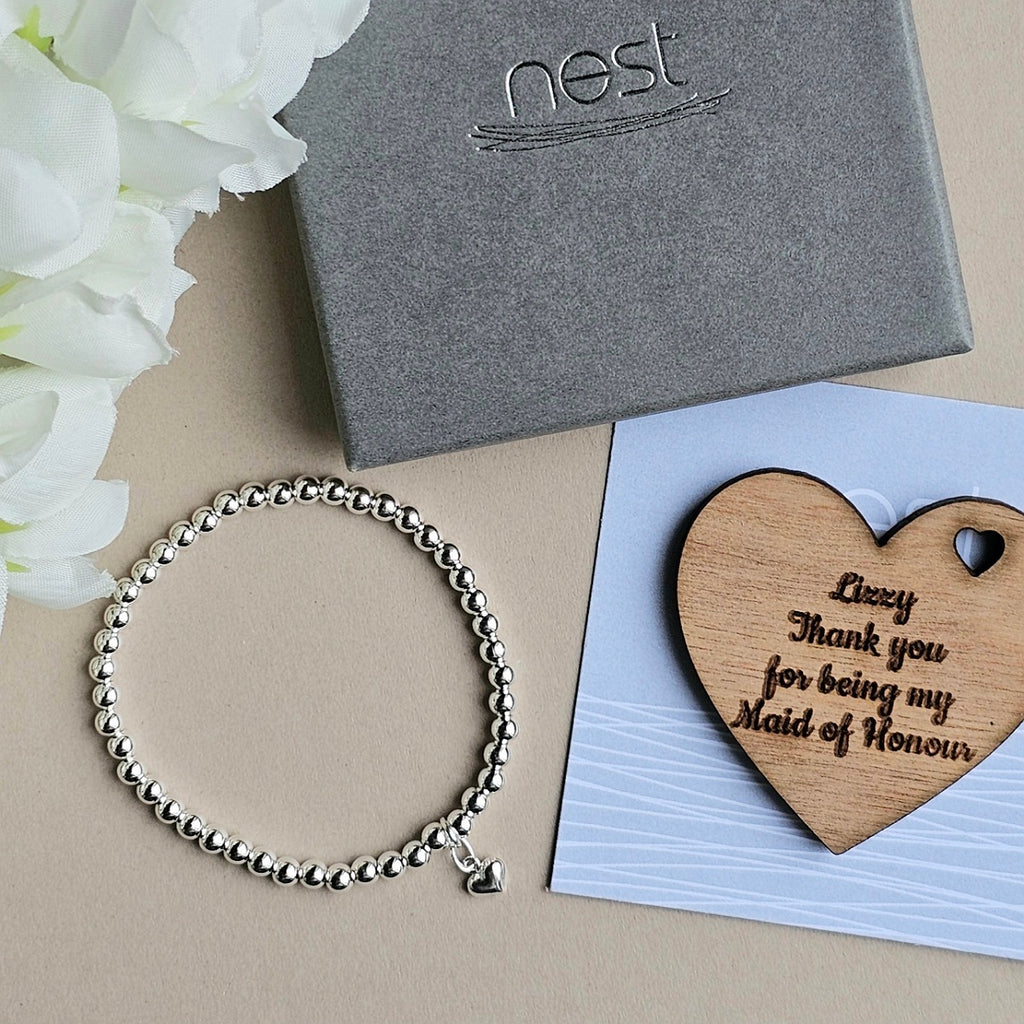 A silver beaded bracelet with a small heart charm is displayed on a beige surface next to a gray Nest gift box, white flowers, and a wooden heart-shaped keepsake engraved with 'Lizzy Thank you for being my Maid of Honour'.