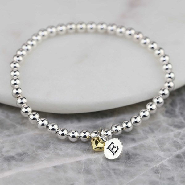 Silver beaded bracelet with a heart-shaped gold charm and a letter 'E' charm on a marble surface.
