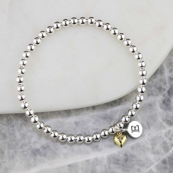 A silver beaded bracelet with two charms, including a gold heart charm, displayed on a white marble slab over a gray marble surface.