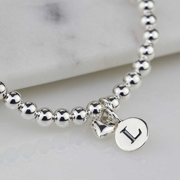A silver beaded bracelet with a small heart charm and a round disc charm engraved with the letter 'L', displayed on a light marble surface.