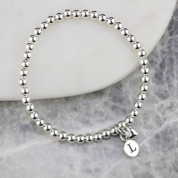 A silver beaded bracelet with a small initial 'L' charm, displayed on a white ceramic plate over a gray marble surface.