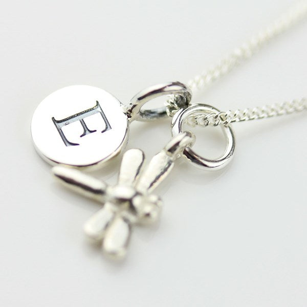 A silver necklace with a round disc charm engraved with a monogram letter 'E', a small dragonfly charm, and a chain on a light gray background.