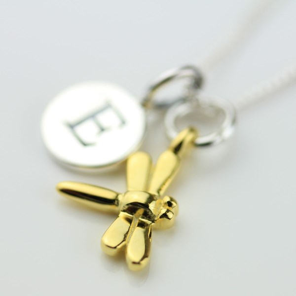 A close-up of a gold dragonfly pendant necklace with a silver round initial disc engraved with the letter 'E' and a silver ring charm on a white chain, set against a plain white background.
