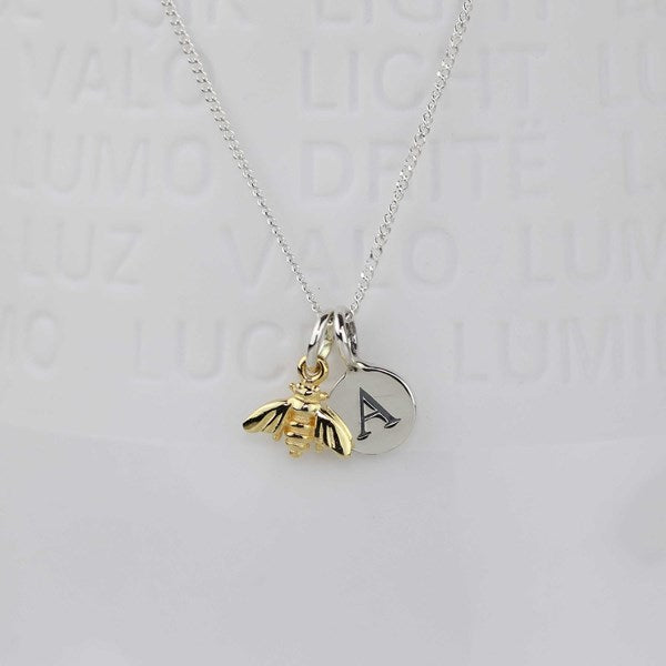 A silver chain necklace featuring a gold bumblebee charm and a round silver disc charm engraved with the letter 'A', displayed against a light gray background with subtle brand text.