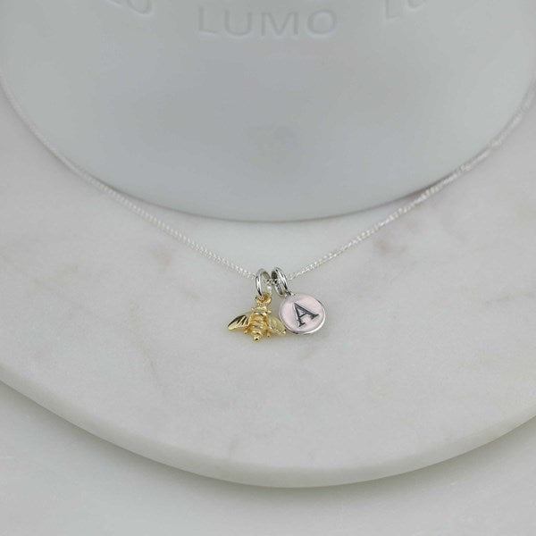A delicate silver chain necklace with a small charm featuring a gold bee and a silver disc engraved with the letter 'A', displayed on a white marble stand.