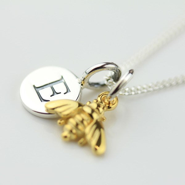 A silver and gold pendant necklace featuring a round silver disc engraved with a letter, a gold bee charm, and a silver lobster clasp on a silver chain, shown on a light gray background.
