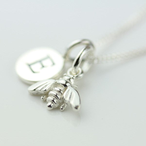 A sterling silver necklace featuring a detailed bee charm, a round engraved initial 'E' disc charm, and a delicate chain on a light gray background.
