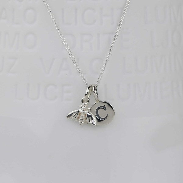 A silver necklace with a delicate chain and a pendant featuring a small bee charm next to a circular disc engraved with the letter 'C', displayed against a light gray background with faint embossed text.