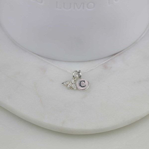 A delicate silver necklace with a small bee charm and a letter 'C' pendant, displayed on a white marble stand.