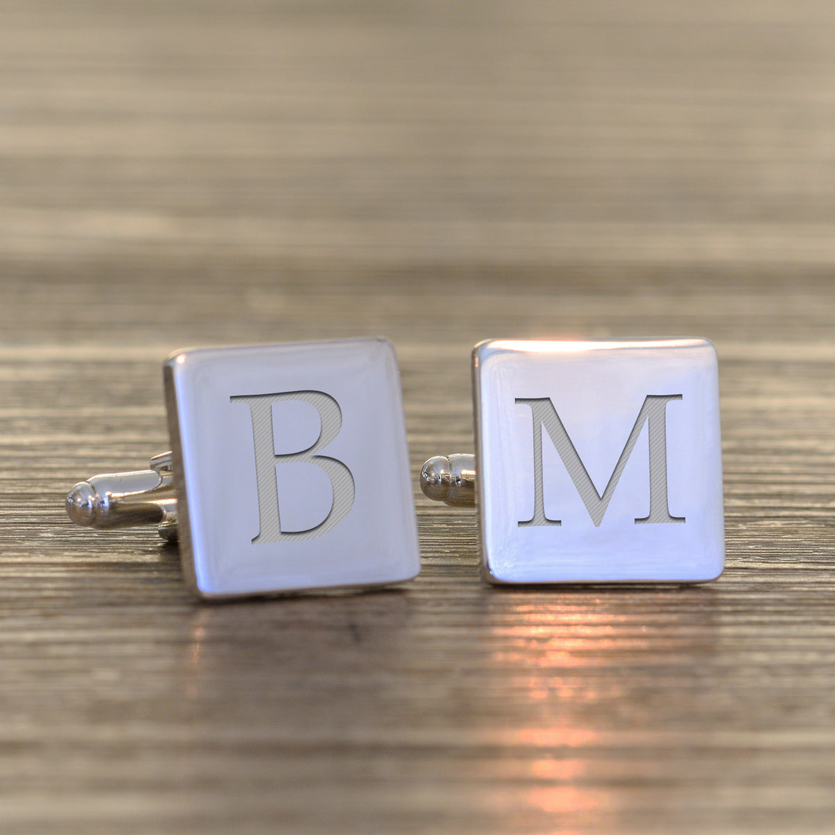 A pair of square silver cufflinks with engraved letters 'B' and 'M' on a wooden surface.