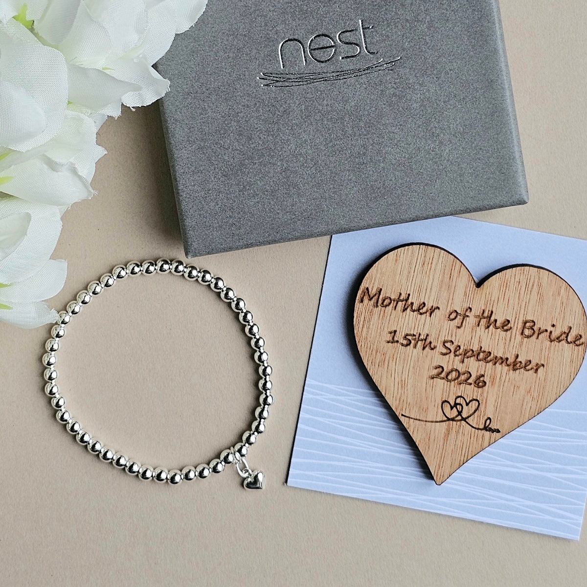 Silver bracelet and wooden heart with engraving on a beige surface with flowers and Nest box.