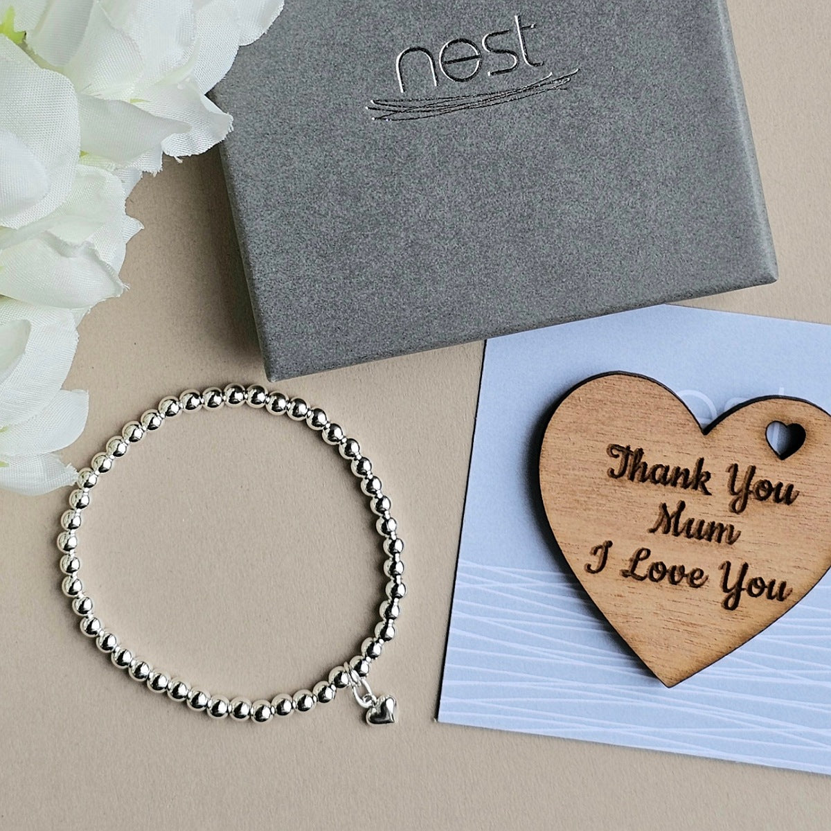 Silver bracelet with heart charm next to a wooden heart-shaped card with 'Thank You Mum I Love You' text, on a beige surface with white flowers.