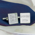 A pair of silver square cufflinks displayed on a dark navy blue fabric over a white dress shirt. One cufflink is engraved with 'Dad 1992' and the other with 'Grandad 2016'.
