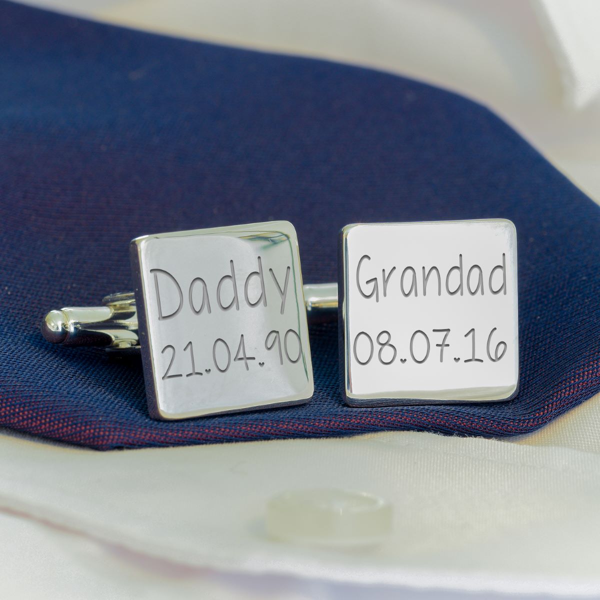A pair of silver square cufflinks displayed on a dark navy blue fabric over a white dress shirt. One cufflink is engraved with 'Daddy 21.04.90' and the other with 'Grandad 08.07.16'.