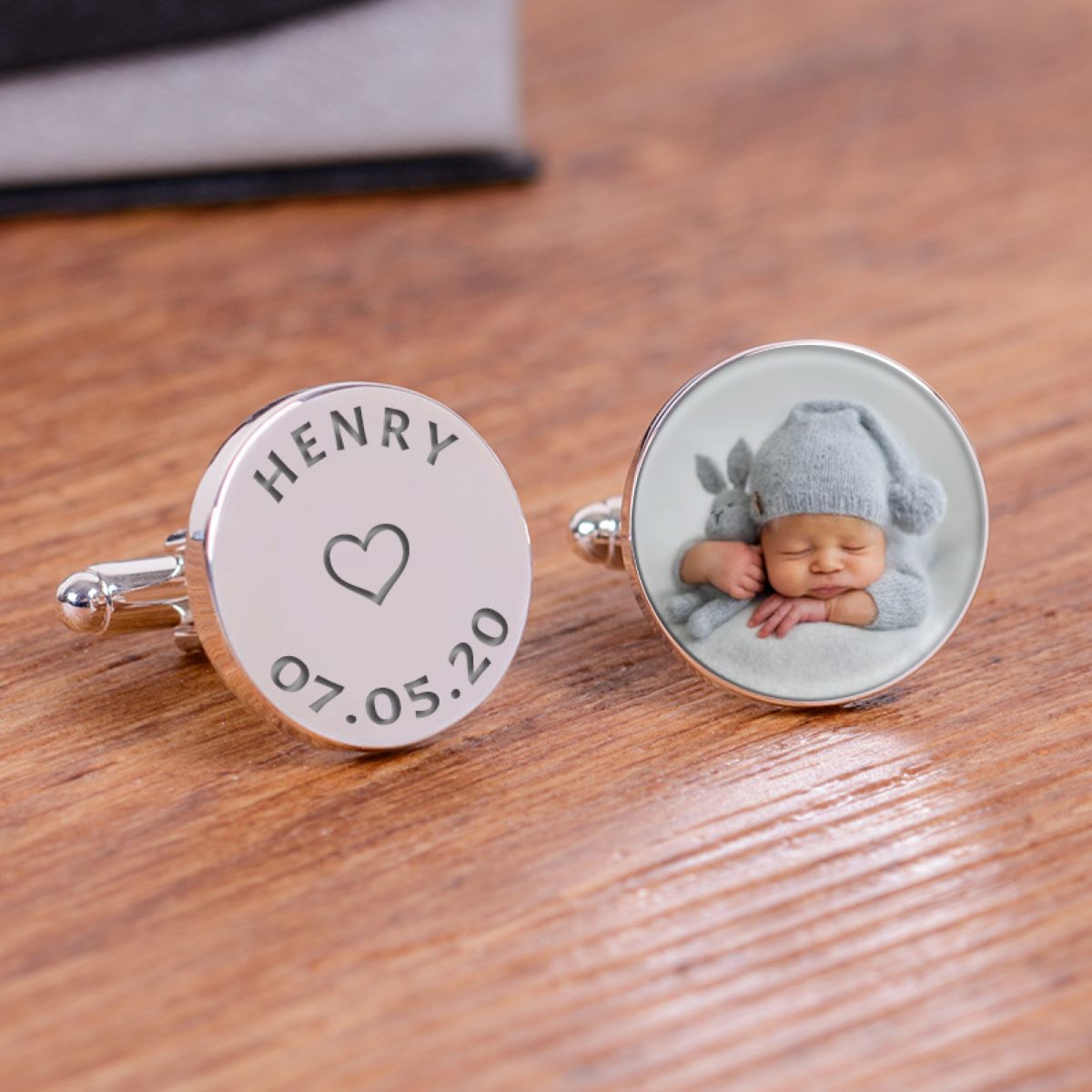 A pair of round silver cufflinks on a wooden tabletop. One cufflink is engraved with the name 'HENRY', a heart, and the date '07.05.20' and the other a photo.