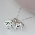 A silver necklace featuring a textured elephant charm and a round engraved disc charm with the letter 'A', displayed on a light pink surface.