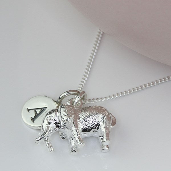 A silver necklace featuring a textured elephant charm and a round engraved disc charm with the letter 'A', displayed on a light pink surface.