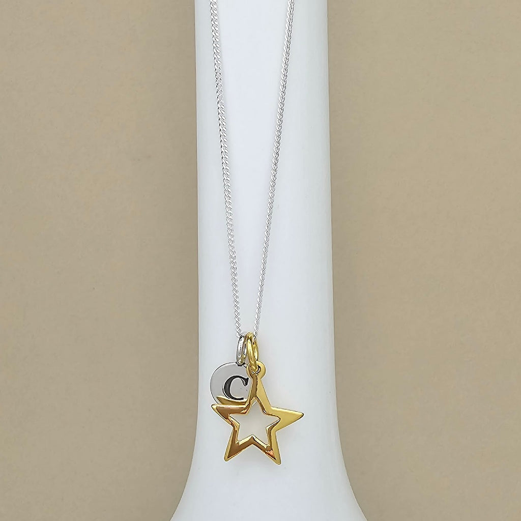 A delicate necklace with a silver chain, featuring a gold star pendant and a small silver disc charm engraved with the letter 'C', displayed on a white jewelry bust against a beige background.