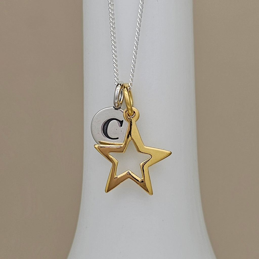 A silver and gold initial necklace featuring a star and crescent moon pendant with the letter 'C', displayed on a white neck stand against a beige background.