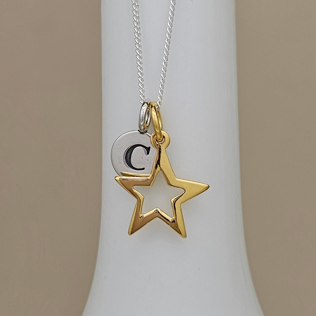 A silver and gold initial necklace featuring a star and crescent moon pendant with the letter 'C', displayed on a white neck stand against a beige background.