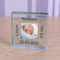 A square personalised clear glass block display on a light wood surface, featuring a photo of a baby and engraved text reading 'William 7lbs 2oz 6.35pm 20.03.2023' on each side of the photo.
