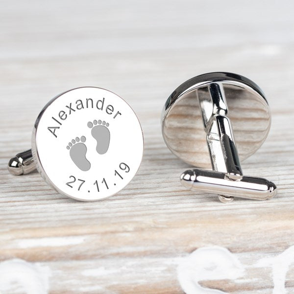 Personalised silver cufflinks on a light wood background. One cufflink is shown from the front with engraved text reading 'Alexander' and baby footprints with the date '27.11.19'.