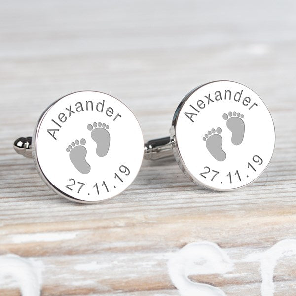 A pair of round silver cufflinks with engraved baby footprints and the name 'Alexander' along with the date '27.11.19.