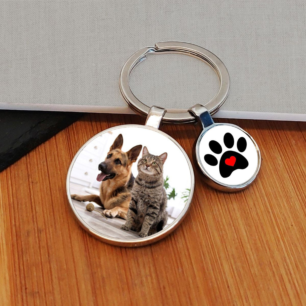 A custom photo keyring with a round metal ring, featuring two charms: one displaying a photo of a cat and dog, and the other showing a black paw print with a red heart on a white background. The keychain is displayed on a wooden surface.