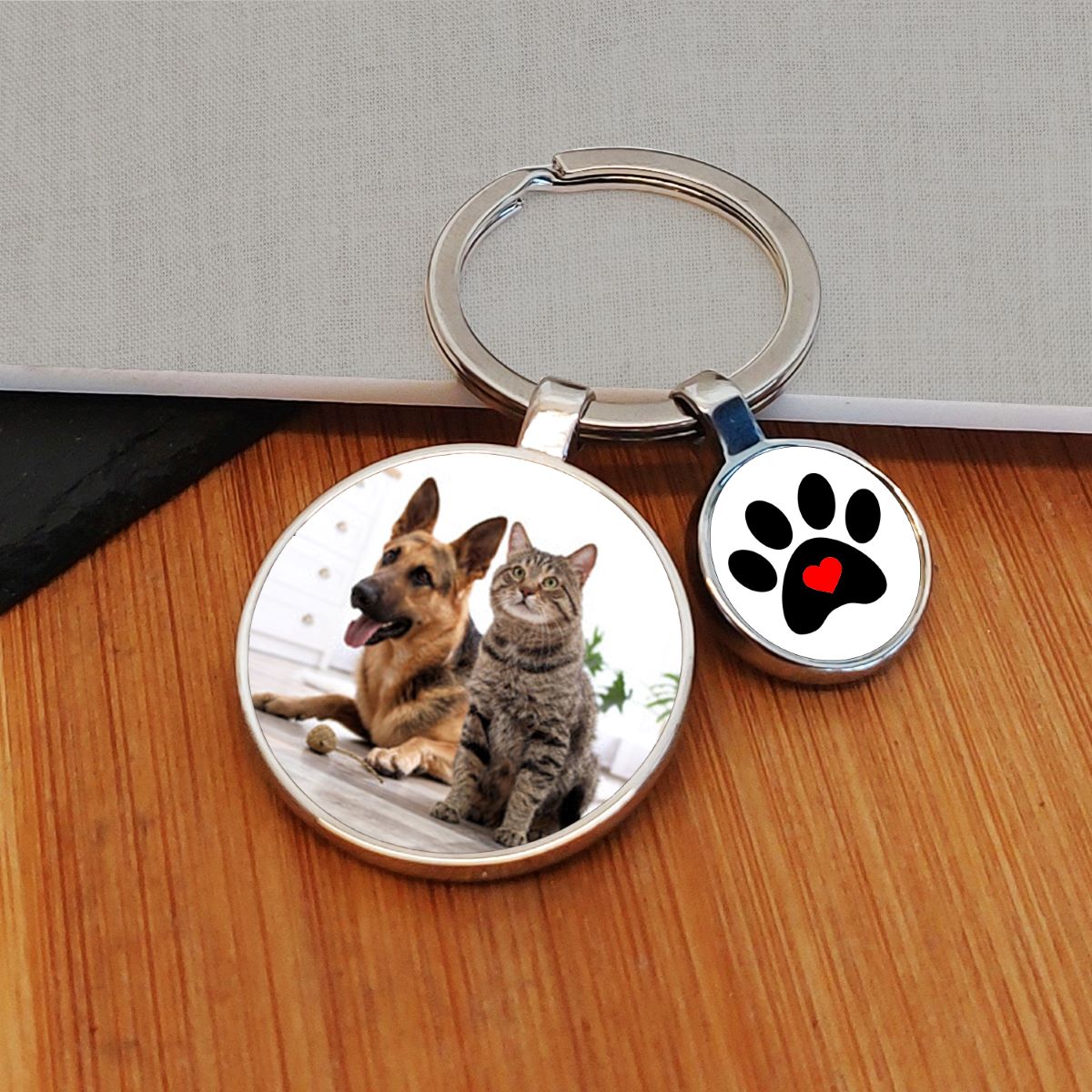 A custom photo keyring with a round metal ring, featuring two charms: one displaying a photo of a cat and dog, and the other showing a black paw print with a red heart on a white background. The keychain is displayed on a wooden surface.