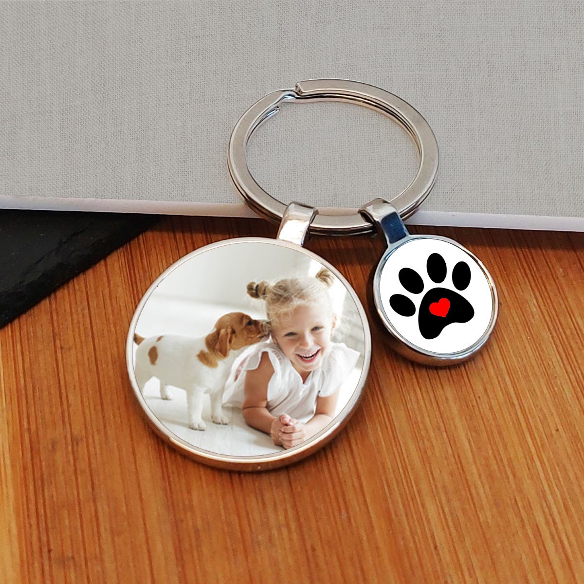 A custom photo keyring with a round metal ring, featuring two charms: one displaying a photo of a young girl and a puppy, and the other showing a black paw print with a red heart on a white background. The keychain is displayed on a wooden surface.