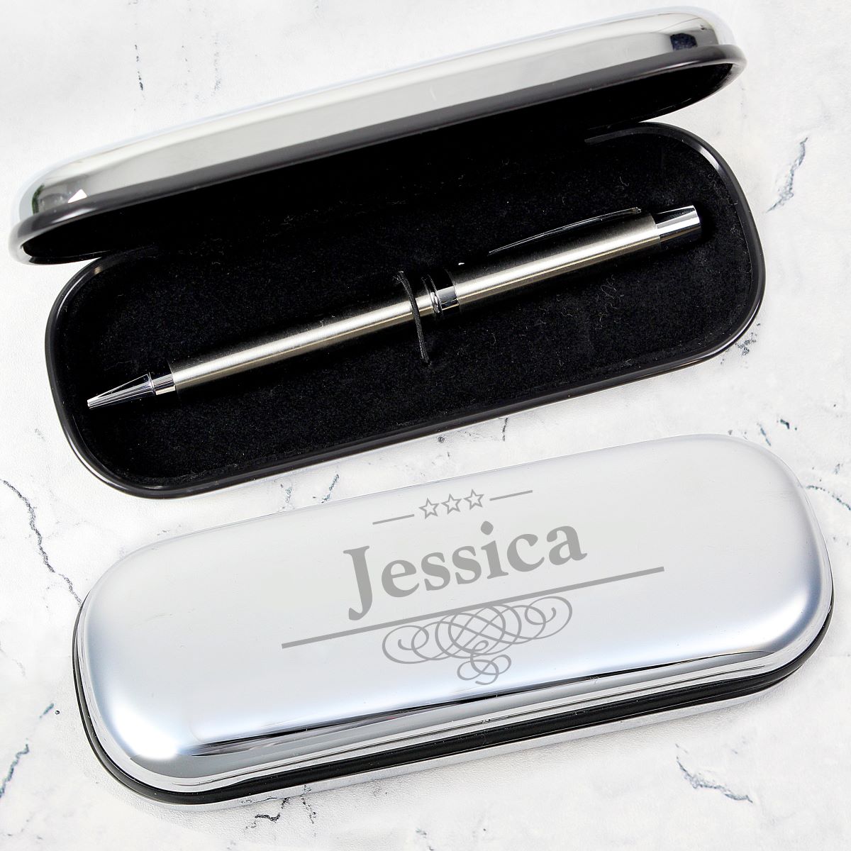 A silver metal pen displayed inside an open black case, with a closed matching case in front engraved with the name 'Jessica' and decorative stars and swirls, set on a light marble surface.