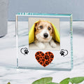 A clear glass square photo frame with a custom pet portrait of a dog, decorated with a black paw print and a small orange heart with black paw prints on the front, set on a marble surface.