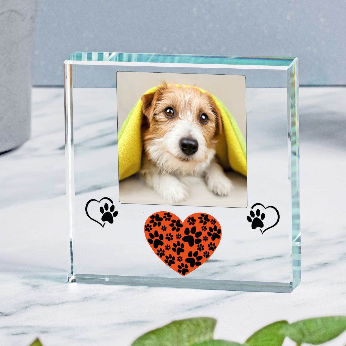 A clear glass square photo frame with a custom pet portrait of a dog, decorated with a black paw print and a small orange heart with black paw prints on the front, set on a marble surface.