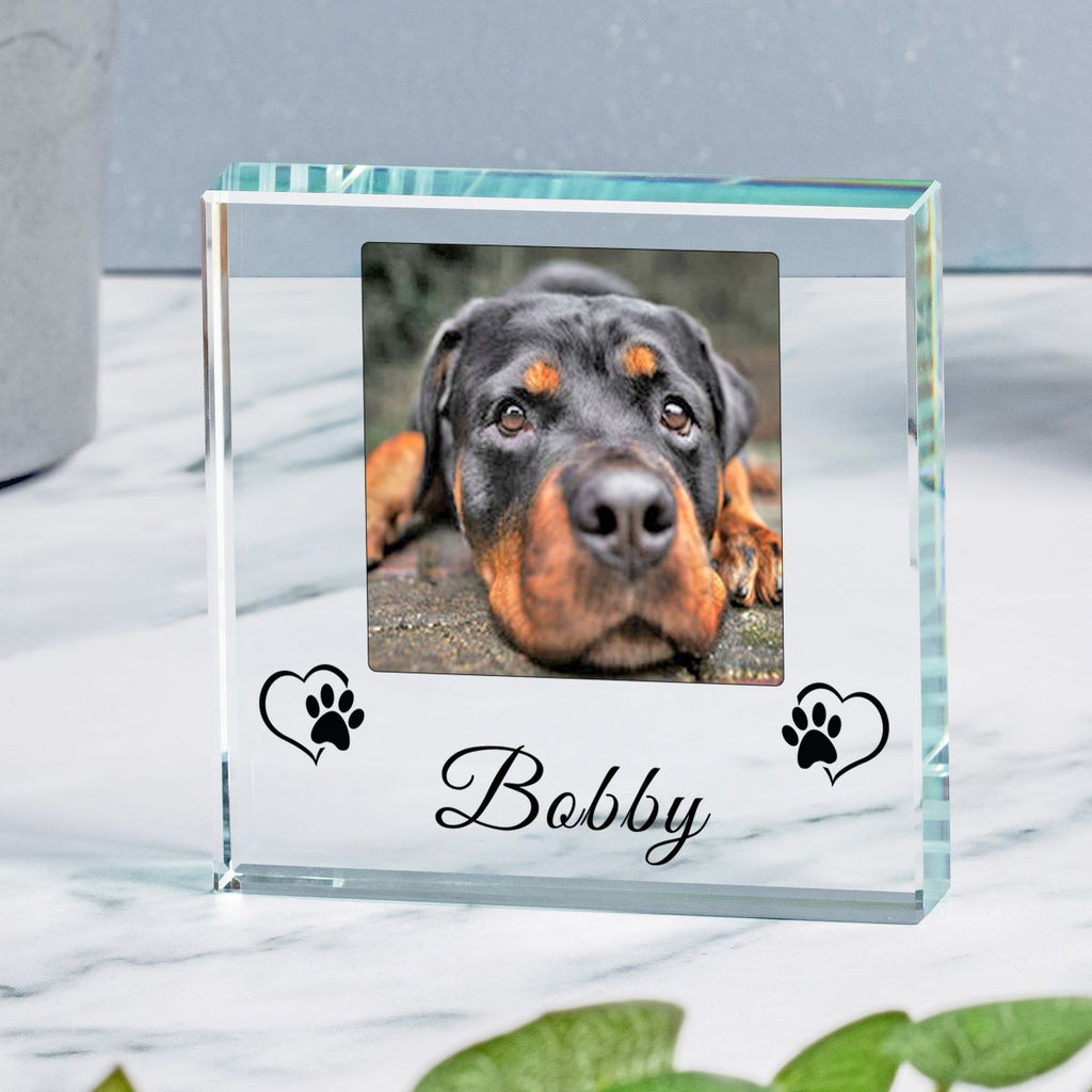 A clear glass square photo frame with a custom photo of a dog, featuring the name 'Bobby' and black paw print heart designs on the front, displayed on a white marble surface.