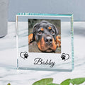 A clear glass square photo frame with a custom photo of a dog, featuring the name 'Bobby' and black paw print heart designs on the front, displayed on a white marble surface.