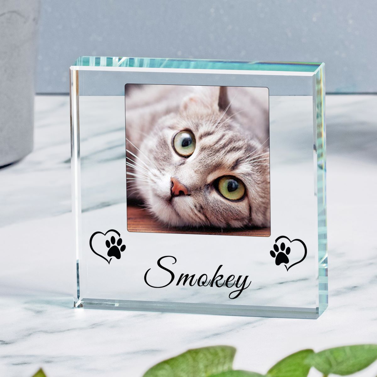 A clear glass square photo frame with a custom photo of a grey tabby cat, featuring the name 'Smokey' and black paw print heart designs on the front, displayed on a white marble surface.