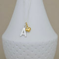 A silver chain necklace with a silver letter 'A' pendant and a small gold heart charm, displayed on a white textured vase against a beige background.