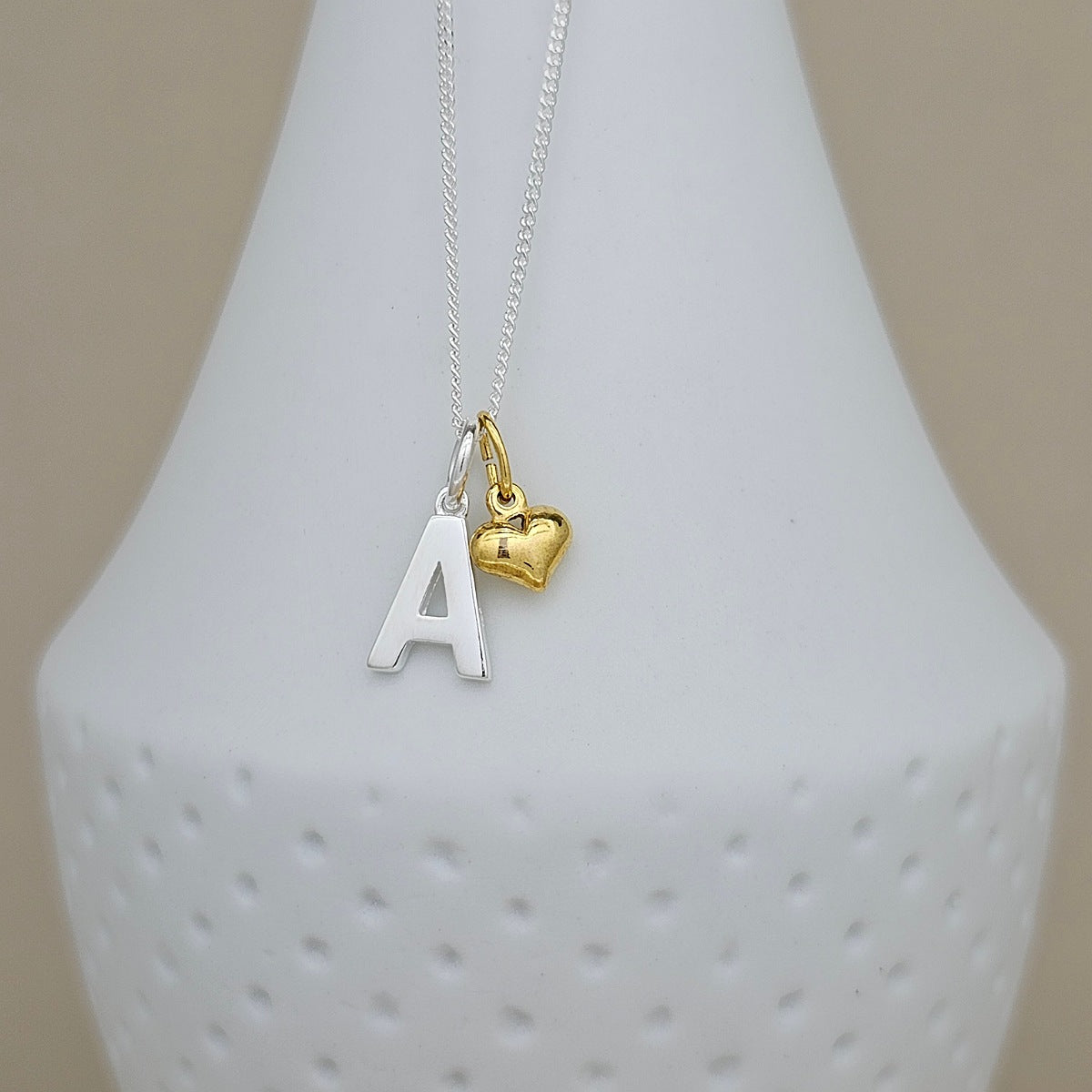 A silver chain necklace with a silver letter 'A' pendant and a small gold heart charm, displayed on a white textured vase against a beige background.