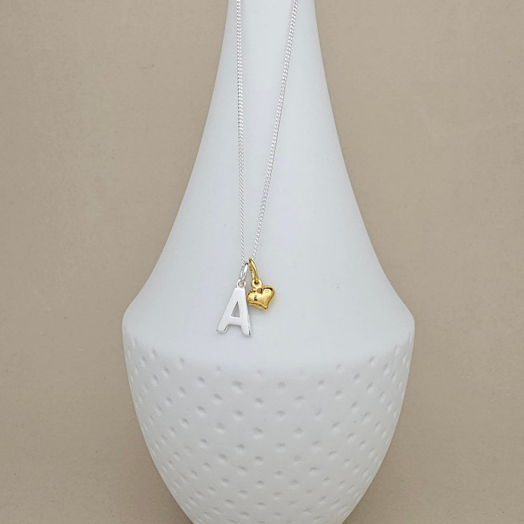 A delicate necklace with a silver-toned chain, featuring a white letter 'A' pendant and a small gold heart charm, displayed on a white textured vase against a beige background.