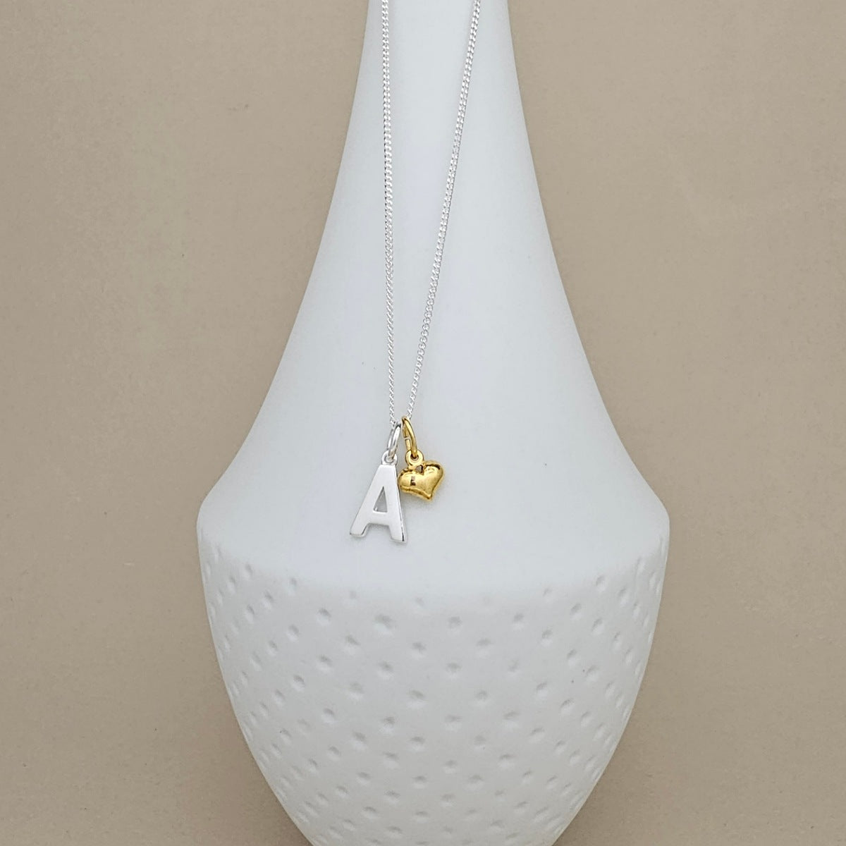 A delicate necklace with a silver-toned chain, featuring a white letter 'A' pendant and a small gold heart charm, displayed on a white textured vase against a beige background.
