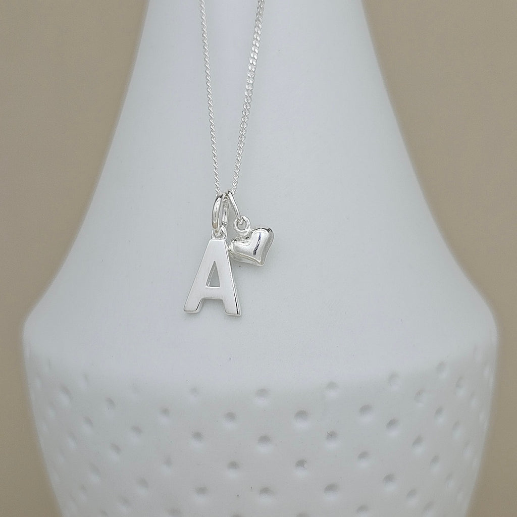 A silver necklace with a letter 'A' initial pendant and a small heart charm, displayed on a white textured ceramic vase against a neutral beige background.