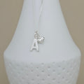 A silver necklace with a letter 'A' initial pendant and a small heart charm, displayed on a white textured ceramic vase against a neutral beige background.
