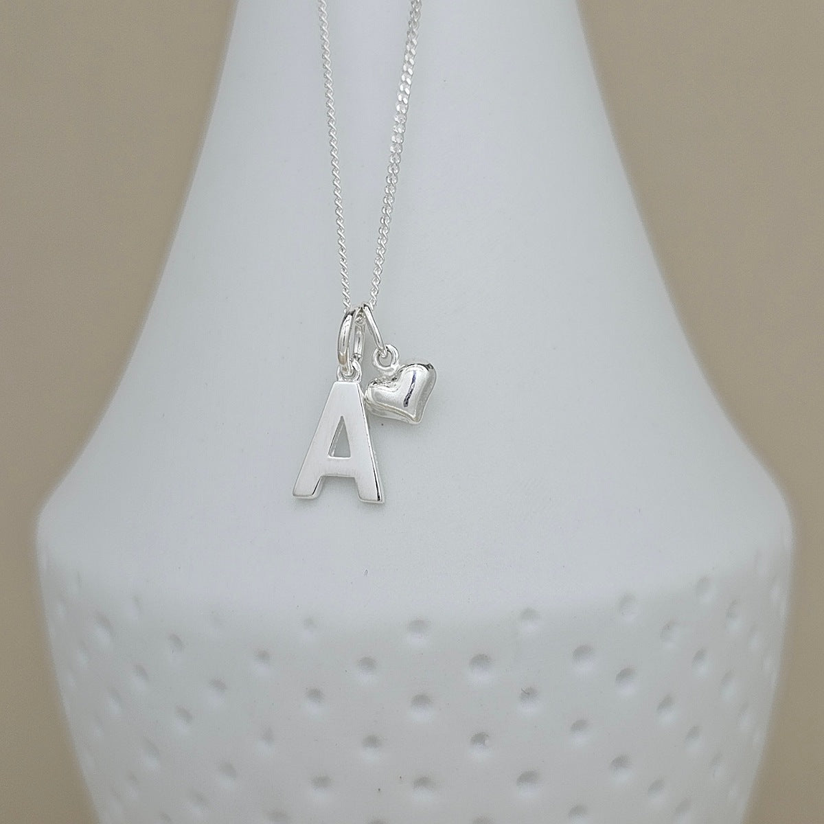 A silver necklace with a letter 'A' initial pendant and a small heart charm, displayed on a white textured ceramic vase against a neutral beige background.