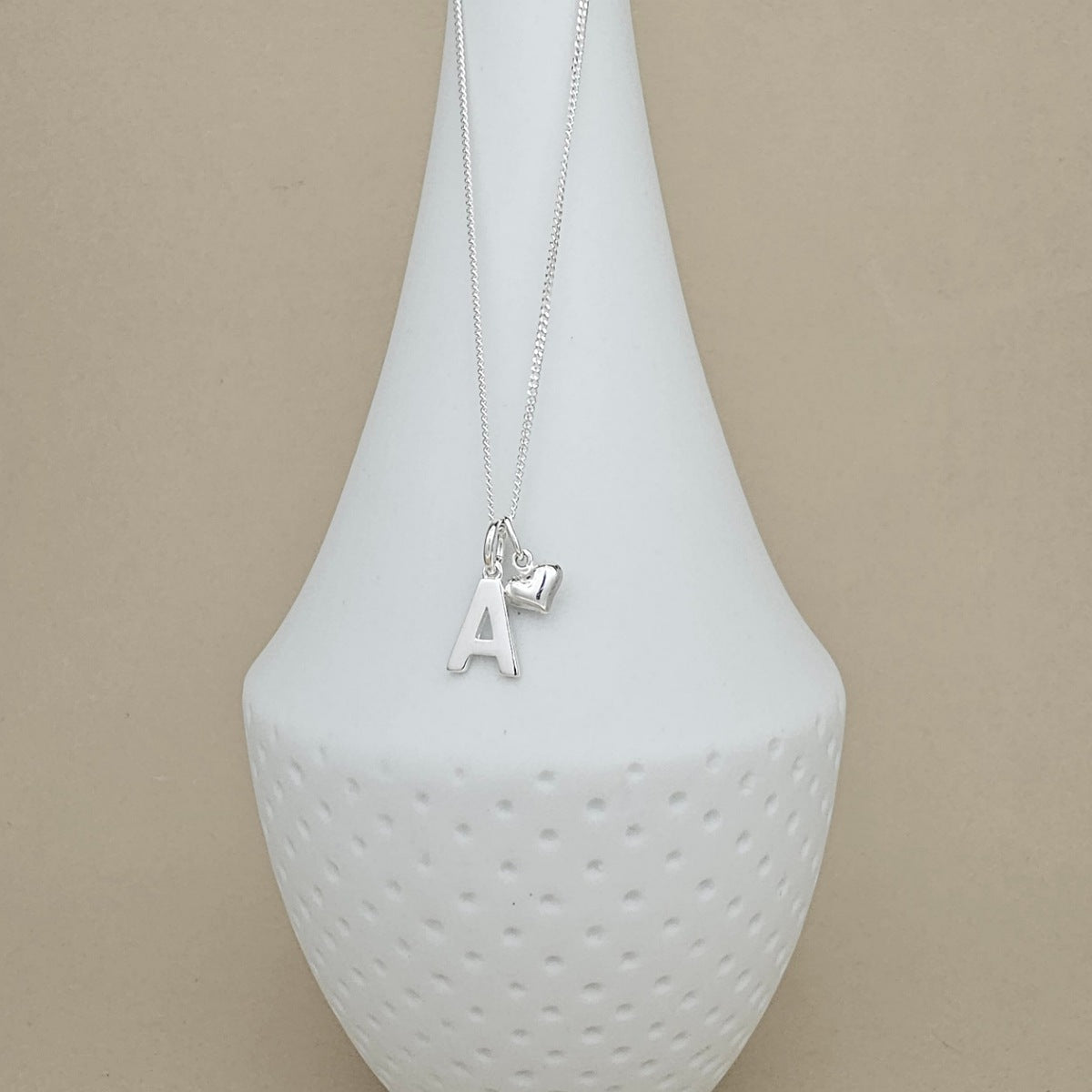 A silver initial necklace with the letter 'A' and a small heart charm, displayed on a white textured ceramic vase against a beige background.
