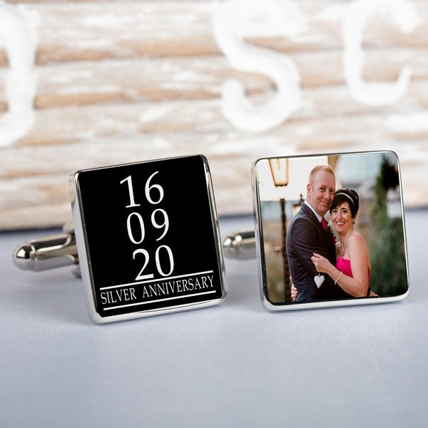 A pair of square silver-toned cufflinks displayed on a light wood surface. One cufflink shows personalised white text on a black background reading '10 09 20 SILVER ANNIVERSARY', the other cufflink displays a photo.