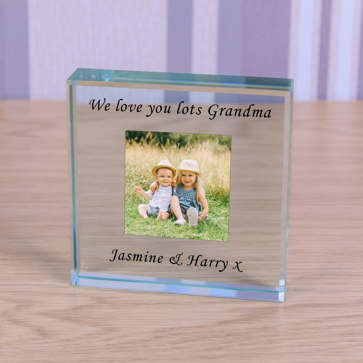 A personalized clear glass photo block plaque engraved with 'We love you lots Grandma' and 'Jasmine & Harry x', featuring a custom photo of two children sitting on grass, displayed on a wooden tabletop.