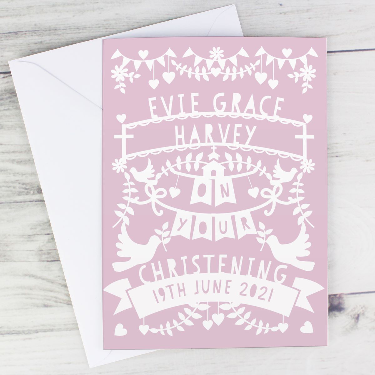 A personalized pink Christening greeting card with white printed decorative text and illustrations, shown with a white envelope on a light wood surface.
