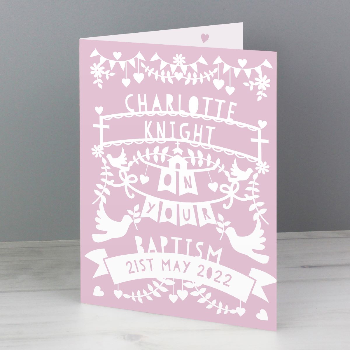A pink personalized baptism greeting card with white cutout-style decorative text and illustrations, standing upright on a light surface against a gray background.
