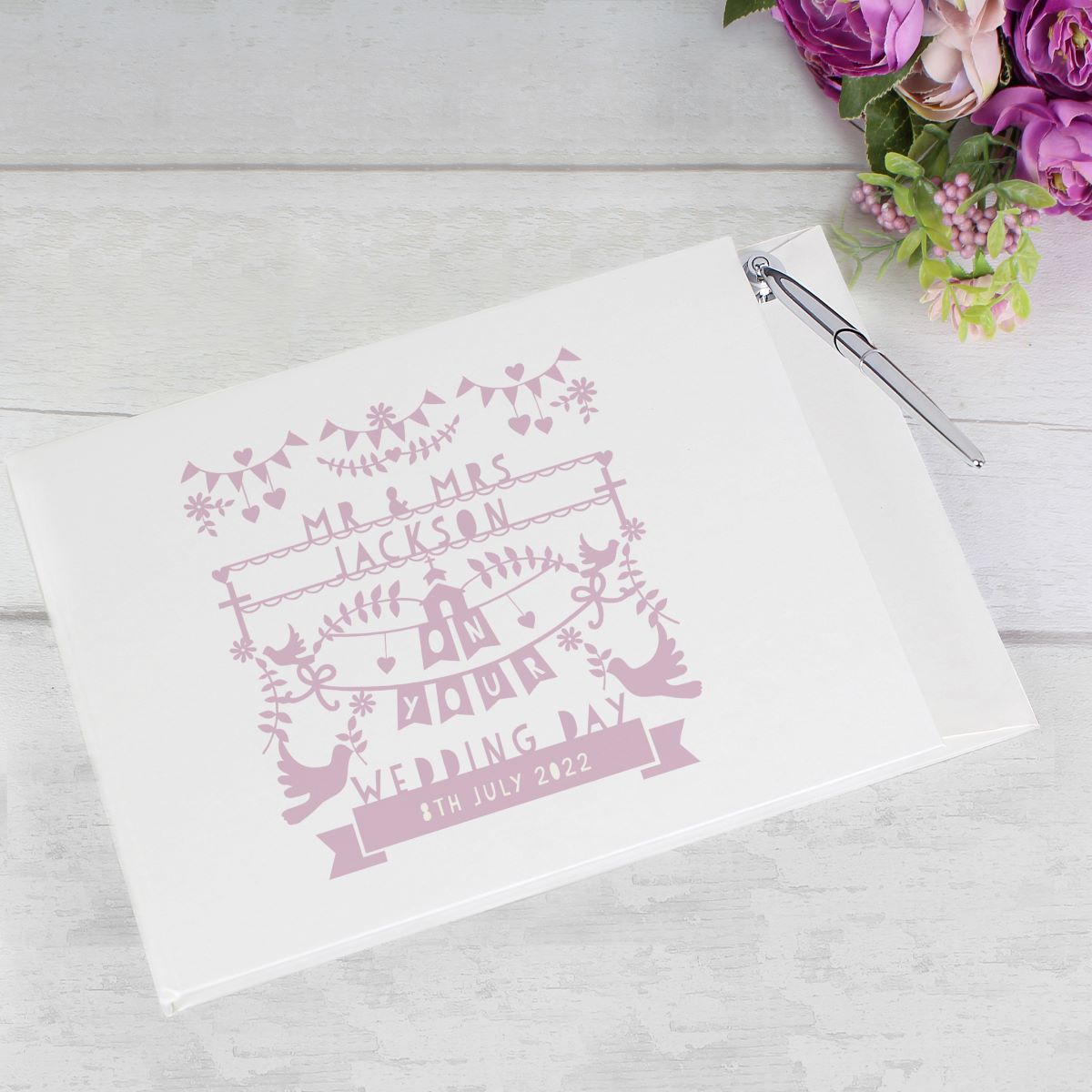 A white wedding guest book with pink personalized text and decorative illustrations, shown on a light wood surface with a silver pen and purple flowers nearby.