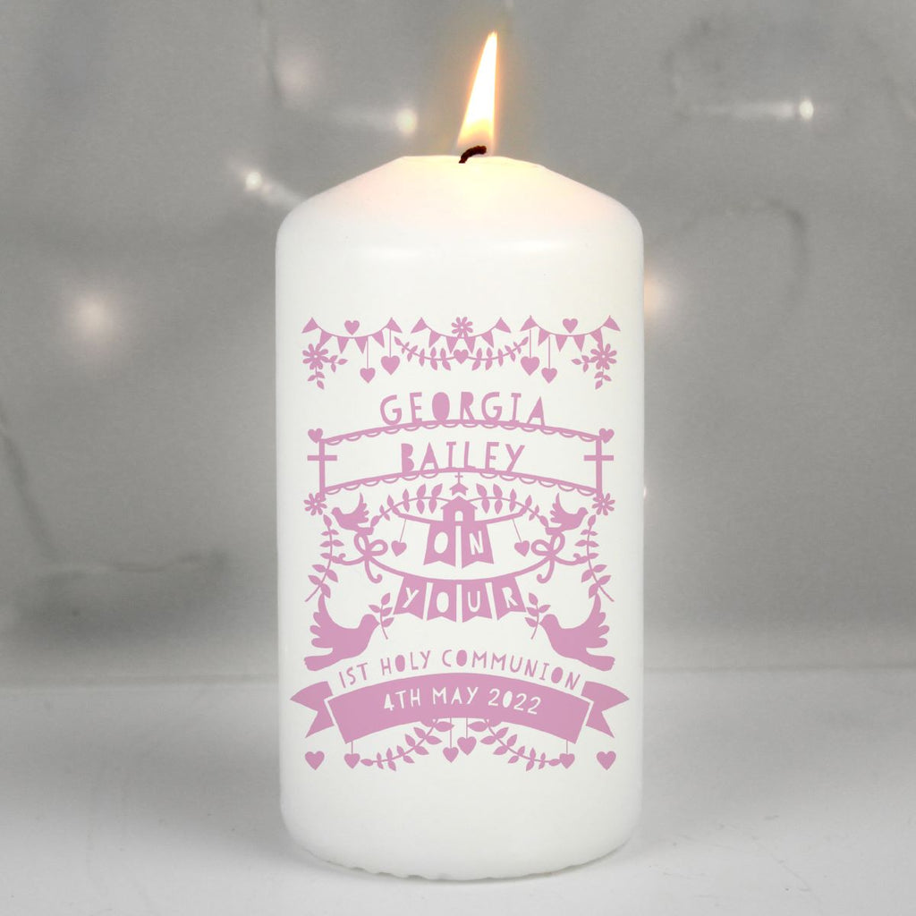 A white pillar candle with a lit flame, featuring pink personalized text and decorative illustrations on the front. The text reads 'GEORGIA BAILEY ON YOUR 1st HOLY COMMUNION 4TH MAY 2022'.